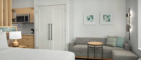 A hotel room with a bed, a gray sofa, a small round table, a microwave in the kitchen, and two framed artworks on the wall.