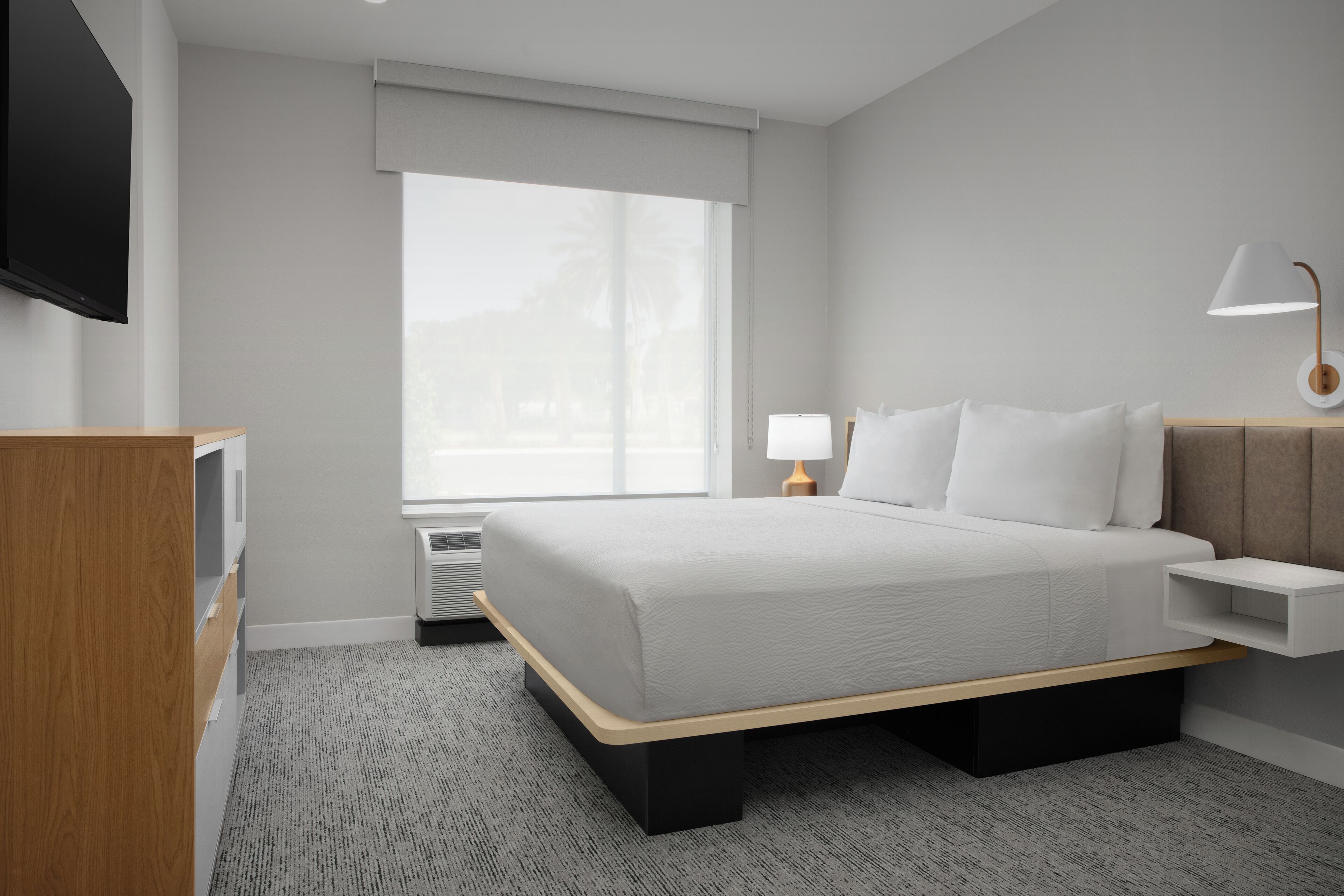 A modern hotel room with a large bed, a TV, a bedside table with a lamp, and a window with a view of palm trees.