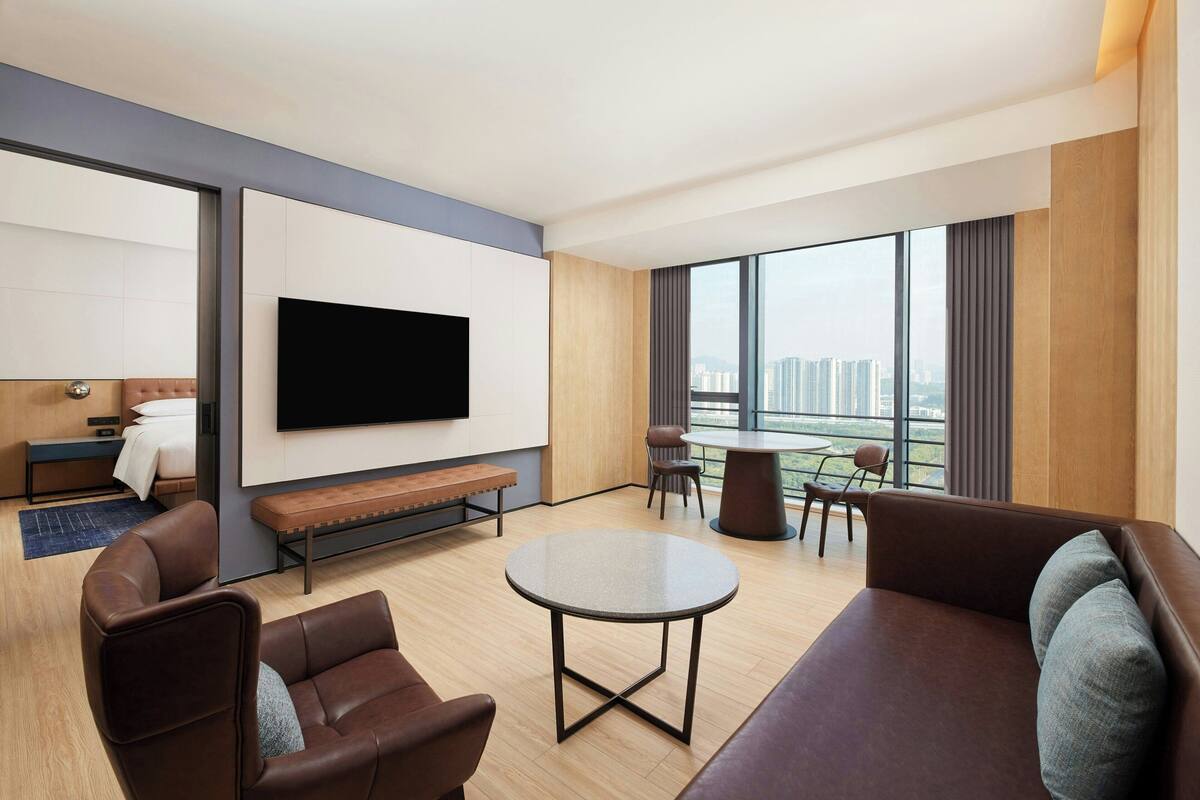A modern hotel room with a large TV, comfortable seating, a balcony with a view of the city, and a wooden floor.