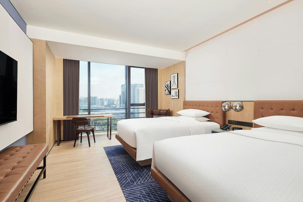 A hotel room with two beds, a desk, a chair, a TV, and a view of the city.