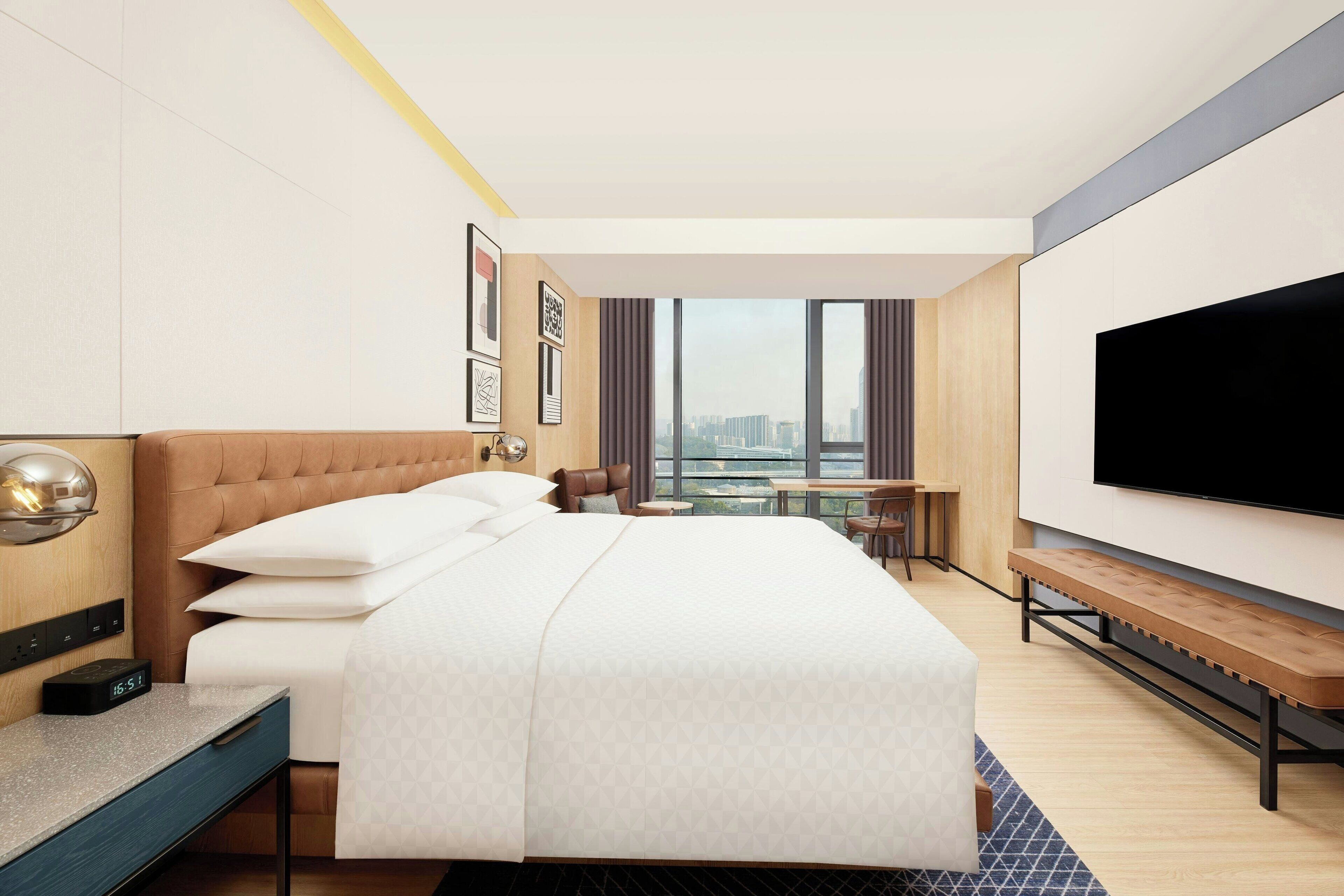 Une chambre d’hôtel moderne dotée d’un grand lit, d’un bureau, d’une télévision à écran plat et offrant une vue sur le paysage urbain.
