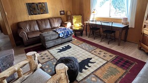 A cozy living room with a brown leather sofa, a wooden coffee table, and a dining table with chairs. There is a large window with curtains, a lamp, and a rug with animal prints.