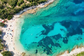 Vue aérienne d’une plage aux eaux turquoise et limpides, avec une côte rocheuse et une zone de plage sablonneuse parsemée de parasols et de personnes.