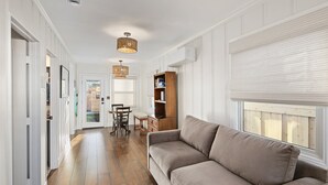 Un pasillo con piso de madera, un sofá color beige, una mesa de comedor con sillas y una estantería.