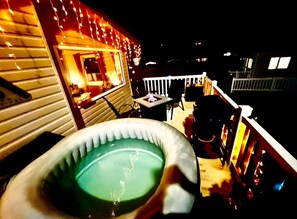 Un jacuzzi en un balcón con vista al cielo nocturno y un edificio iluminado con luces de cadena.
