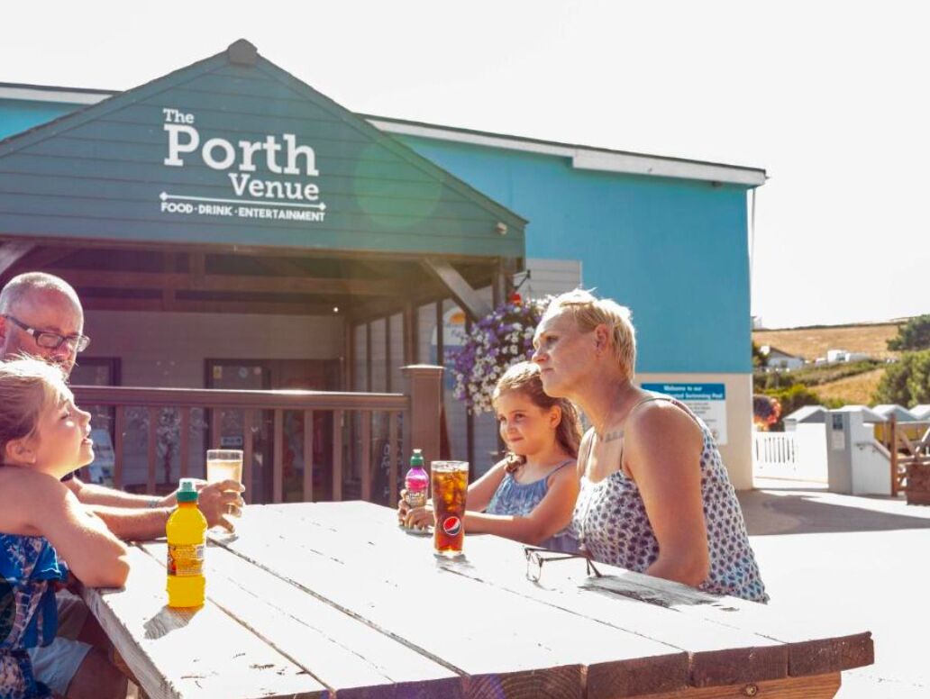 A family at a wooden table outside a venue named "The Porth Venue" with a sign indicating it offers food, drink, and entertainment.
