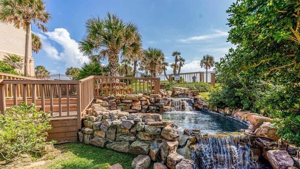 Un jardin avec une petite cascade, une terrasse en bois et des palmiers.