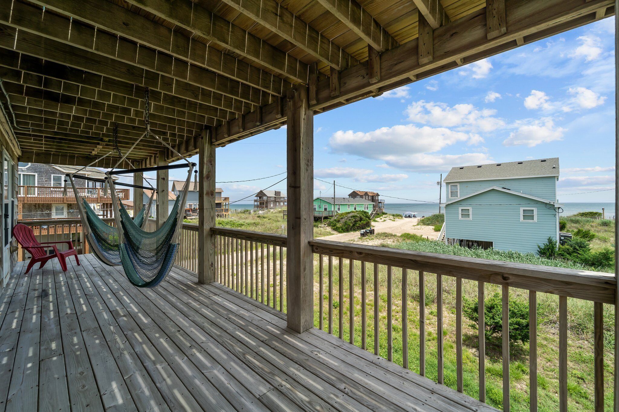 A wooden deck with a hammock and a red chair, overlooking a beach with houses and a clear sky.