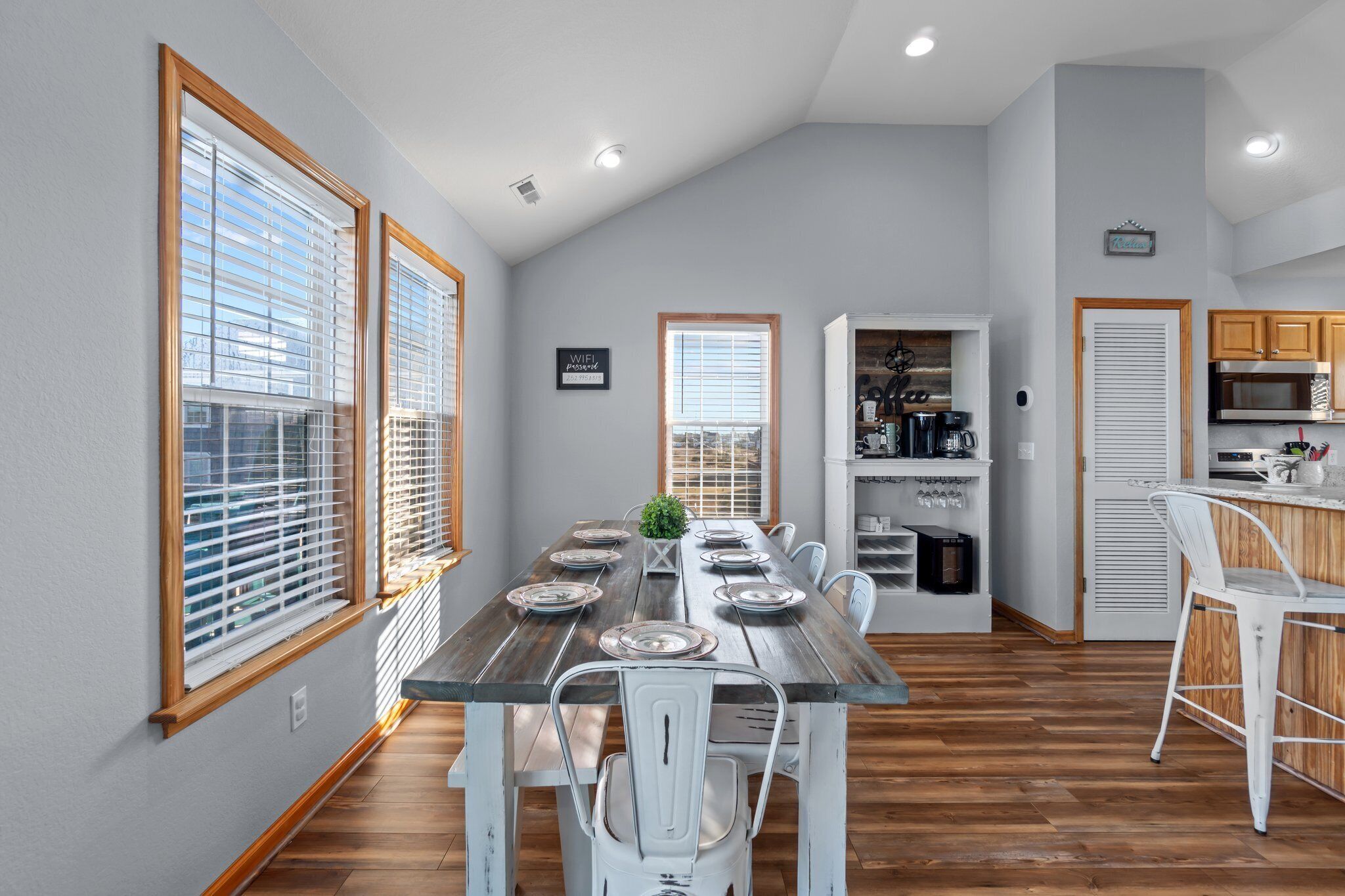 A dining area with a wooden table set for six, white chairs, and a kitchen in the background with wooden cabinets and a microwave.