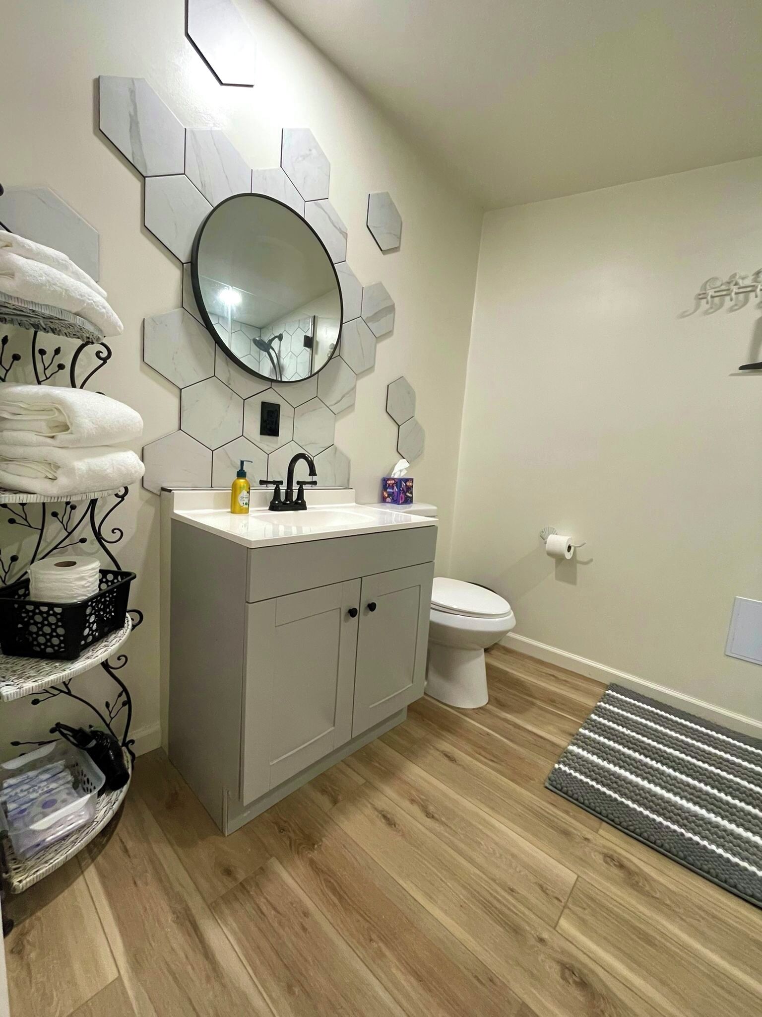 Een moderne badkamer met een grijze wastafel, een ronde spiegel en een toilet. Handdoeken liggen op een plank, er is een zwarte mand en een gestreepte mat op de vloer.