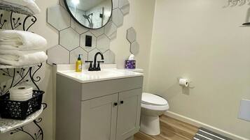 Een moderne badkamer met een grijze wastafel, een ronde spiegel en een toilet. Handdoeken liggen op een plank, er is een zwarte mand en een gestreepte mat op de vloer.