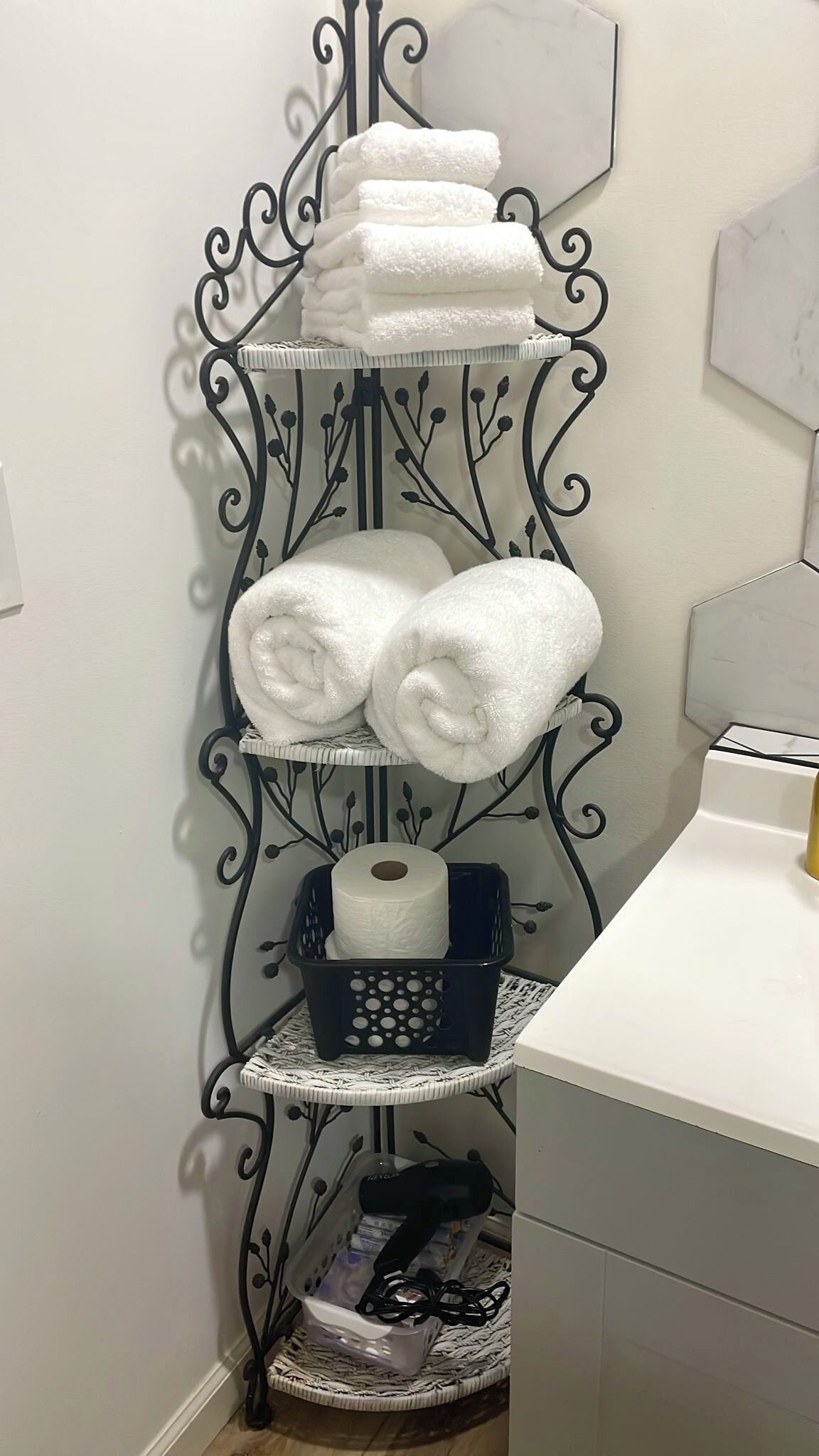 A corner bathroom with a black metal towel rack holding three folded white towels, a black basket with a roll of toilet paper, and a white sink.