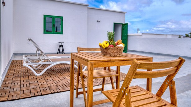 Una mesa de madera con sillas y una cesta de mimbre llena de fruta en una terraza en la azotea, con un edificio blanco al fondo.