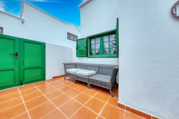 Un pequeño espacio al aire libre amurallado, con un sofá de mimbre, una puerta verde y una ventana con contraventanas verdes.