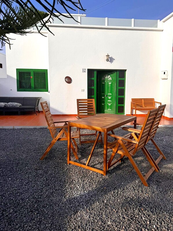 Un área de estar al aire libre con una mesa y sillas de madera, una puerta verde, y una pared blanca con una ventana y un timbre.