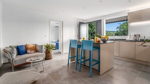 A modern kitchen with a central island, blue bar stools, and a view of greenery outside. - Lounge Appart A refined, discreet, carefully designed space (VAUCLIN)
