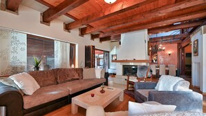 Ein gemütliches Wohnzimmer mit einem braunen Ledersofa, einem grauen Sessel und einem weißen, flauschigen Hocker. In der Mitte steht ein Holztisch mit einer Vase und einer roten Kerze. Das Zimmer verfügt über freiliegende Holzbalken an der Decke und einen Kamin im Hintergrund.
