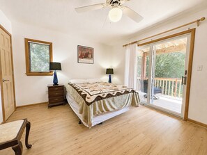 Ein Schlafzimmer mit einem Bett, zwei Nachttischlampen, einem Deckenventilator, einem Fenster mit Jalousien und einer Glastür, die auf eine Terrassenfläche hinausführt.