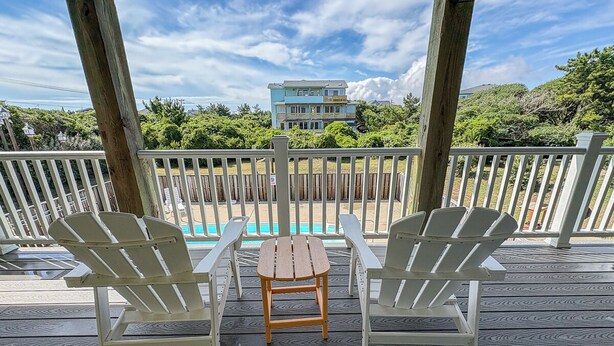 Una terraza de madera con dos sillas Adirondack y una mesita pequeña, con vista a una piscina y un edificio al fondo.