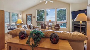 A spacious living room with a large sofa, a ceiling fan, a fireplace, and large windows offering a view of the outdoors. - Wilson's Landing - Enjoy beautiful Sound Views from this Soundfront 4BR home in Duck (Duck)