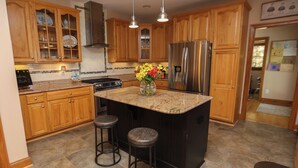 A kitchen with wooden cabinets, a stainless steel refrigerator, a granite countertop, and two stools. - Serendipity on the Sound - Beautiful 4 BR Soundfront Home With Amenities Galore (Southern Shores)