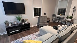 Een moderne woonkamer met een grote grijze modulaire bank, een flatscreen televisie aan de muur, een plafondventilator en een eethoek met een kleine tafel en stoelen.