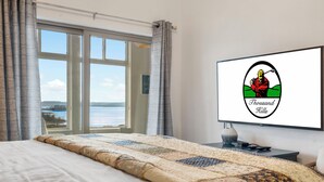 Habitación de hotel con una cama grande, un televisor sobre un mueble y vista al mar a través de un ventanal amplio.