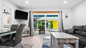 Ein modernes Wohnzimmer mit einer Couch, einem Esstisch und einem Fernseher. Ein Balkon bietet Blick auf einen Pool, und eine Wand ist strahlend gelb.