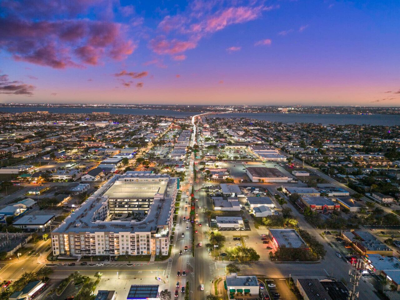 An aerial view of a cityscape at dusk with a prominent road running through the center, surrounded by buildings and a body of water in the background.