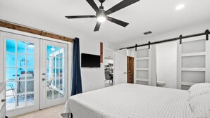 Ein Schlafzimmer mit einem Bett, einem Deckenventilator, einer Glastür, die auf einen Balkon führt, und einem an der Wand befestigten Fernseher.