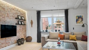 A modern living room with a brick wall, a flat-screen TV, a sofa with cushions, a coffee table with glasses and fruit, and a window with curtains. - Cozy home in Thyborøn with WiFi (Thyborøn)