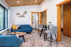 Uma sala de estar moderna com um sofá azul, uma mesa de centro redonda e uma mesa de jantar com cadeiras. Há prateleiras de madeira com objetos decorativos e uma janela grande.