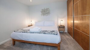 A neatly made bed with white linens and a floral-patterned blanket, a wooden bedside table with a lamp, and a wooden wardrobe with a geometric wall decoration. - Meadow View Nest - Charming Studio in Oakhurst [Sleeps 3] (Oakhurst)