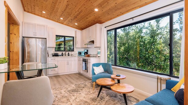 A modern kitchen with white cabinets, a stainless steel refrigerator, and a large window offering a view of greenery. - Meadow View Nest - Charming Studio in Oakhurst [Sleeps 3] (Oakhurst)