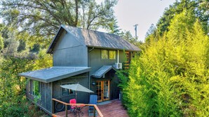 A two-story house with a wooden deck, a covered patio with a table and chairs, and a stone staircase leading to the entrance. - Meadow View Nest - Charming Studio in Oakhurst [Sleeps 3] (Oakhurst)