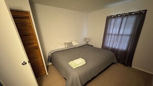 Un dormitorio con cama, mesita de noche, lámpara, ventana con cortinas y puerta que da a otra habitación.