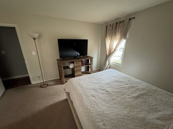 A bedroom with a bed, a TV on a wooden stand, a floor lamp, and a window with curtains.