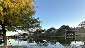 Una vista tranquila del lago con un primer plano de césped, un árbol y un edificio con un porche cubierto al fondo.