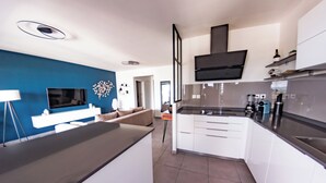 A modern kitchen with white cabinets, a black countertop, and a built-in stove. There is a TV mounted on the wall, and a sofa is visible in the adjacent living area.