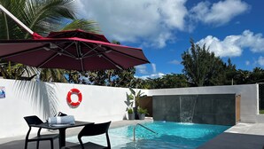 Zona de piscina con mesa y sillas, una sombrilla roja y un salvavidas en la pared.