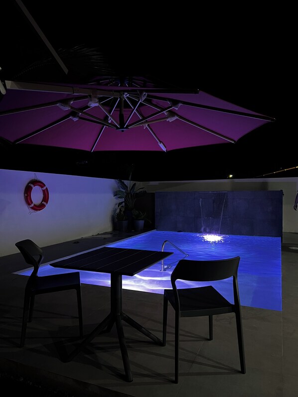 Zona de piscina al aire libre con una mesa, sillas, un paraguas morado y una fuente de agua.