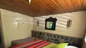 Une chambre avec un plafond en bois, un mur en pierres apparentes, un lit recouvert d’une couverture à rayures et un miroir.