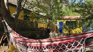 Un hamac suspendu entre deux arbres, un bâtiment jaune avec un mur en pierre et une petite construction bleue.