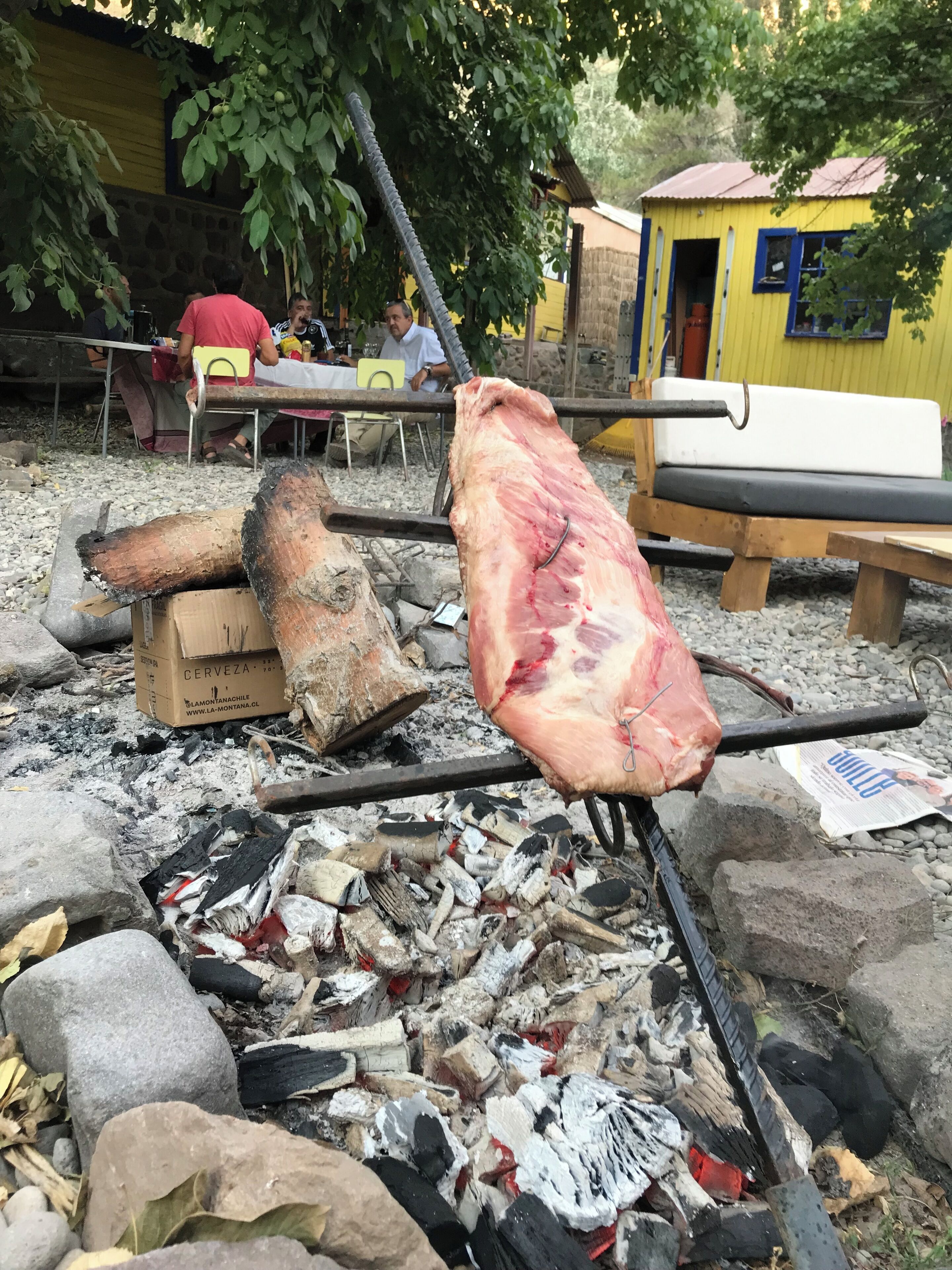 A large piece of meat being cooked over an open fire, with a wooden bench and a yellow building in the background.