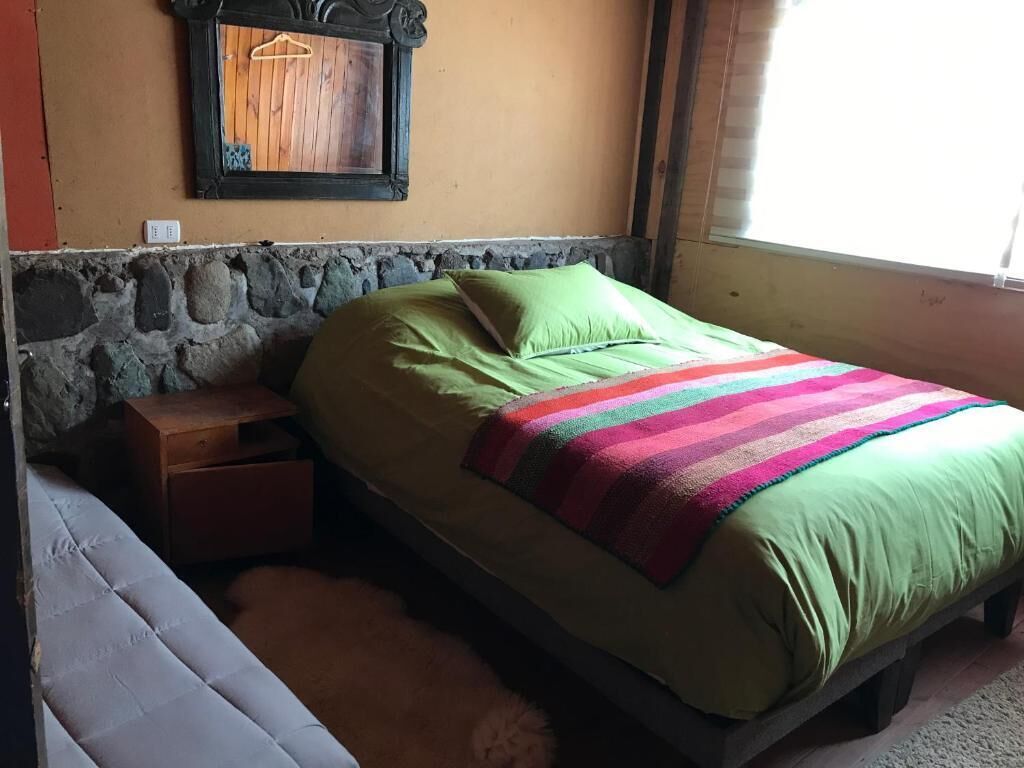 Una cama individual con un almohadón verde y una manta colorida, una mesita de noche de madera y una pared de piedra.