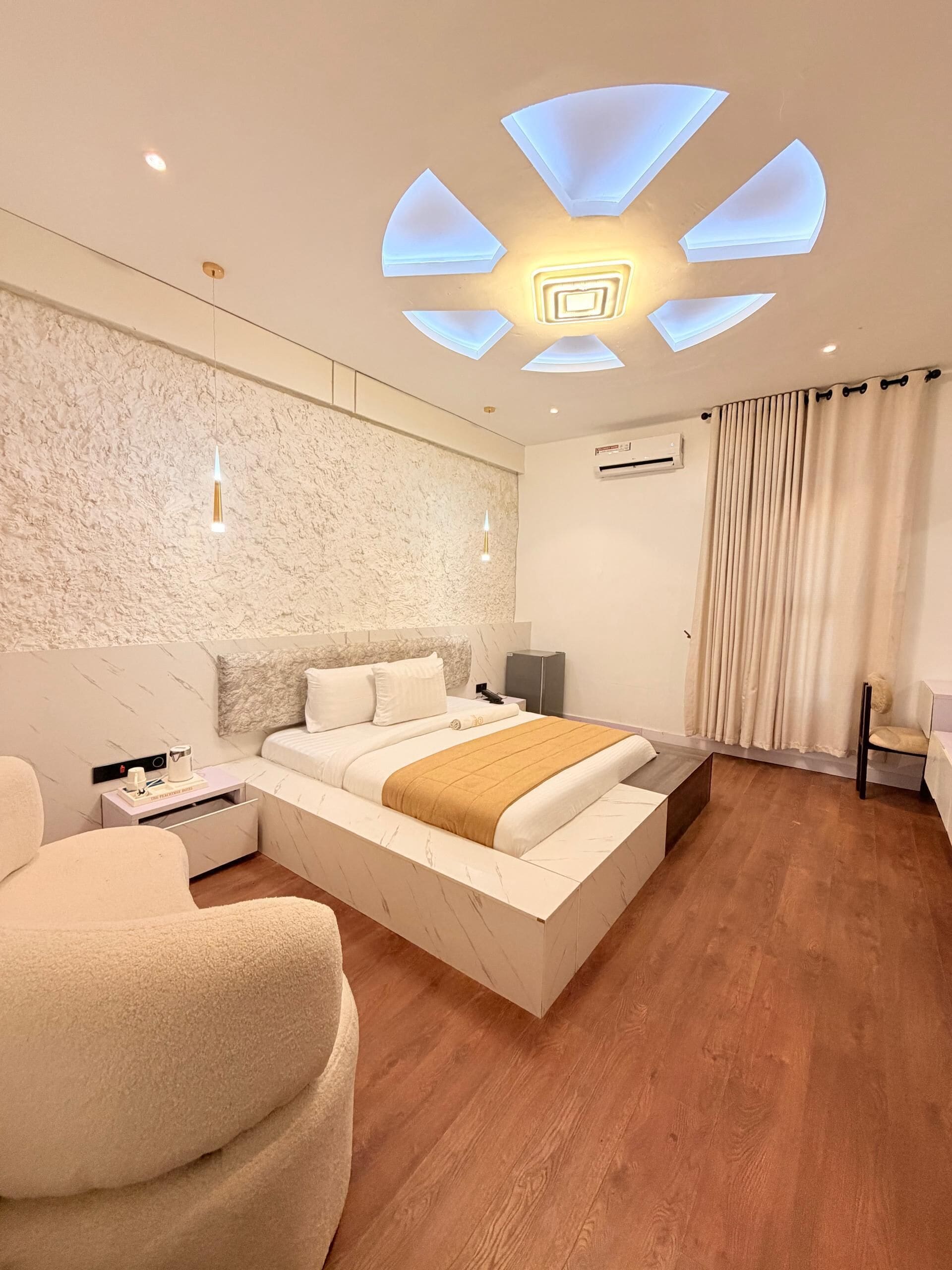 Una habitación de hotel moderna con una cama grande, una mesita de noche, una silla, una lámpara del techo y un equipo de aire acondicionado.