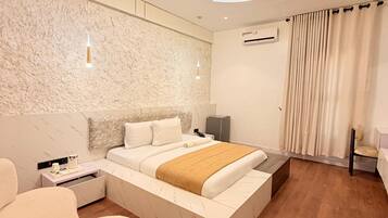 A modern hotel room with a large bed, a bedside table, a chair, a ceiling light fixture, and an air conditioning unit.