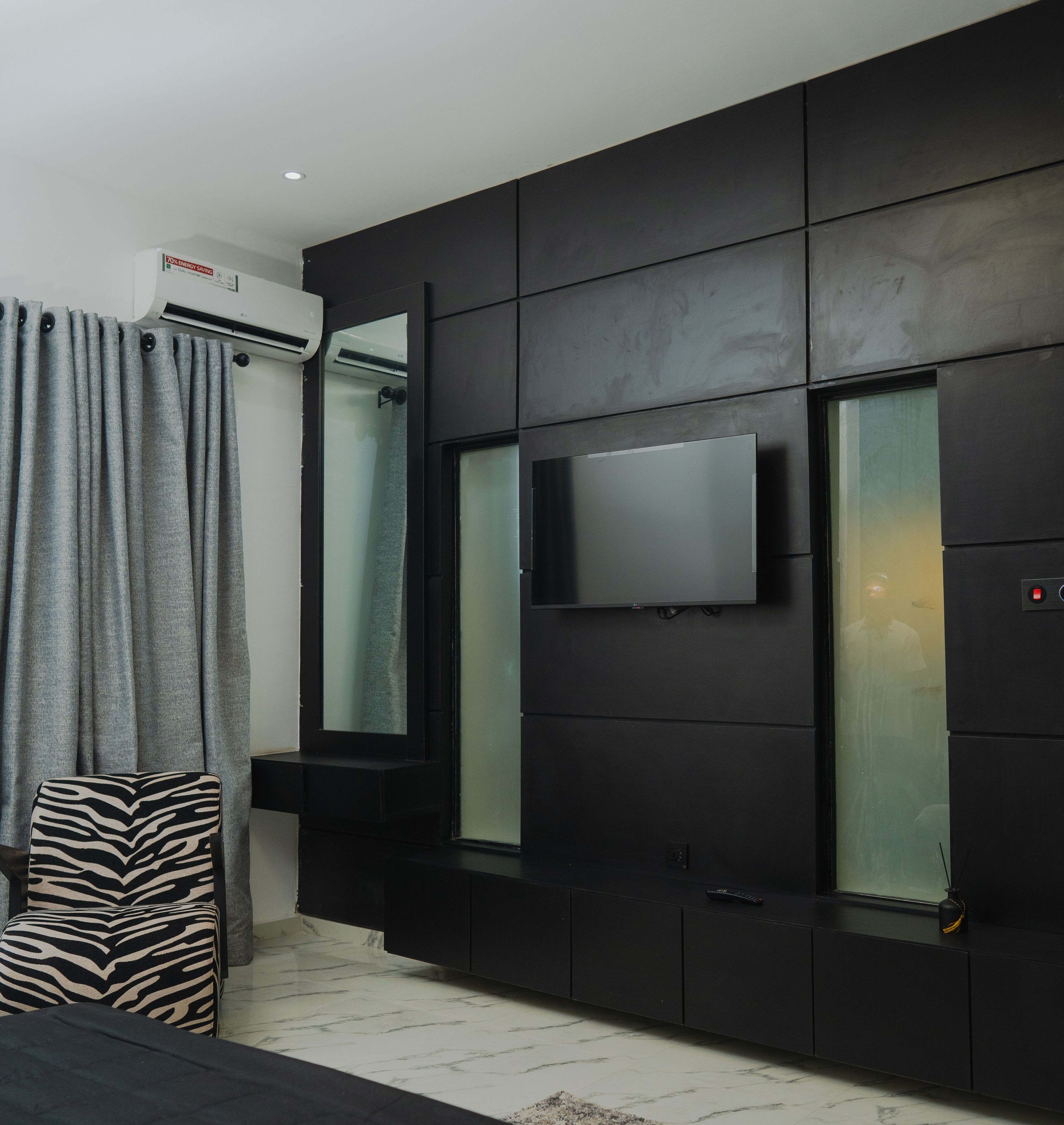 Una sala de estar moderna con un televisor de pantalla plana montado en una pared negra, cortinas grises y un reposapiés con estampado de cebra.