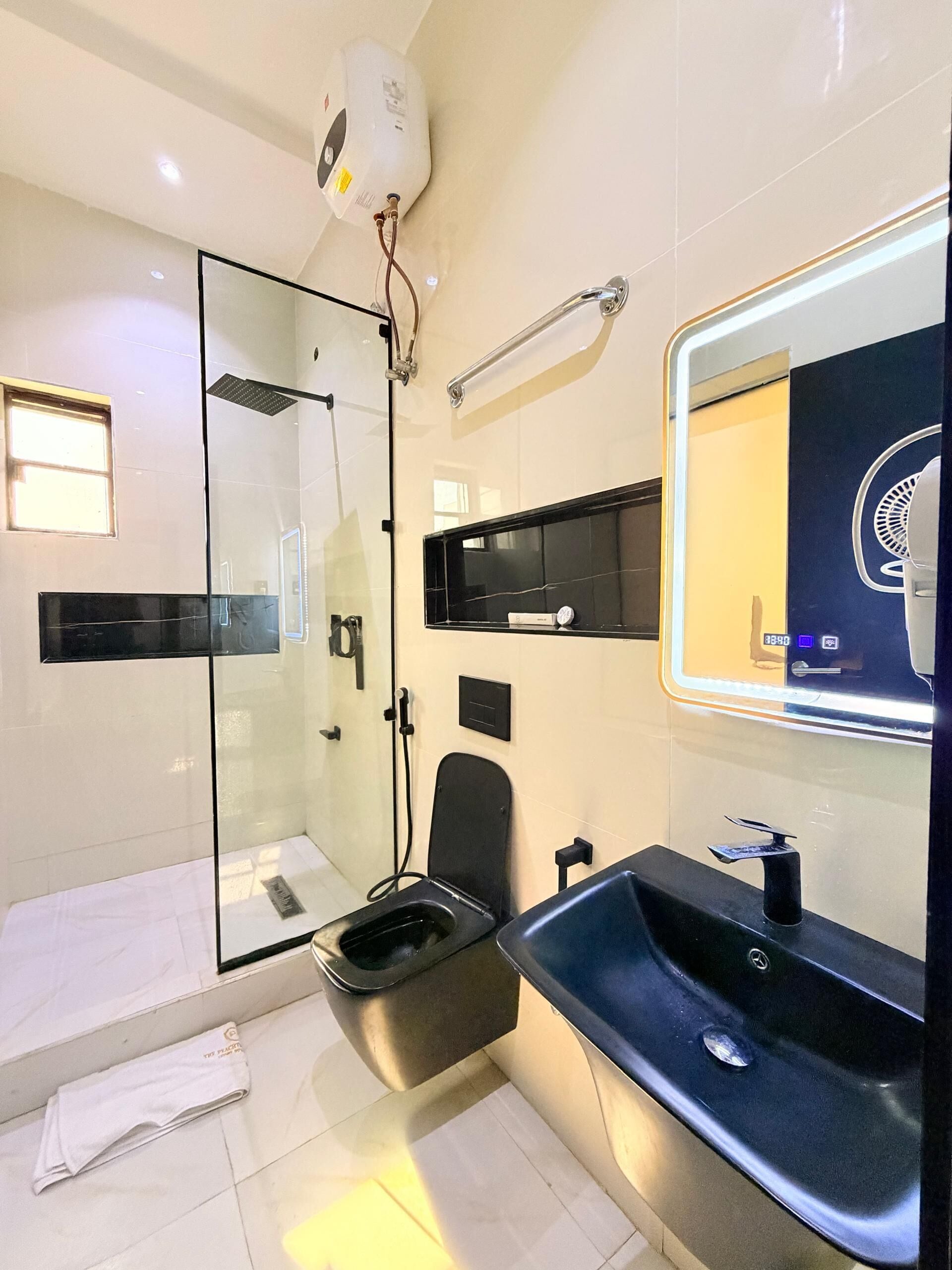 Un baño moderno con cabina de ducha de vidrio, inodoro negro, lavamanos con espejo y una toalla en el suelo.