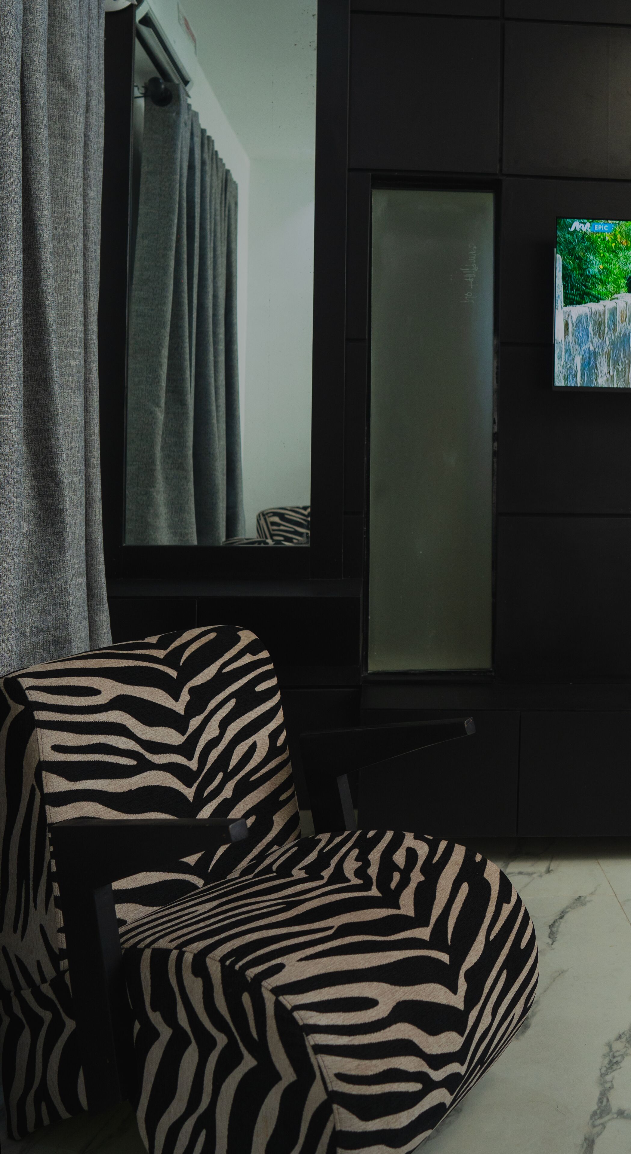 Une pièce avec un fauteuil à motifs zébrés, un meuble sombre et un miroir qui reflète une fenêtre avec des rideaux.